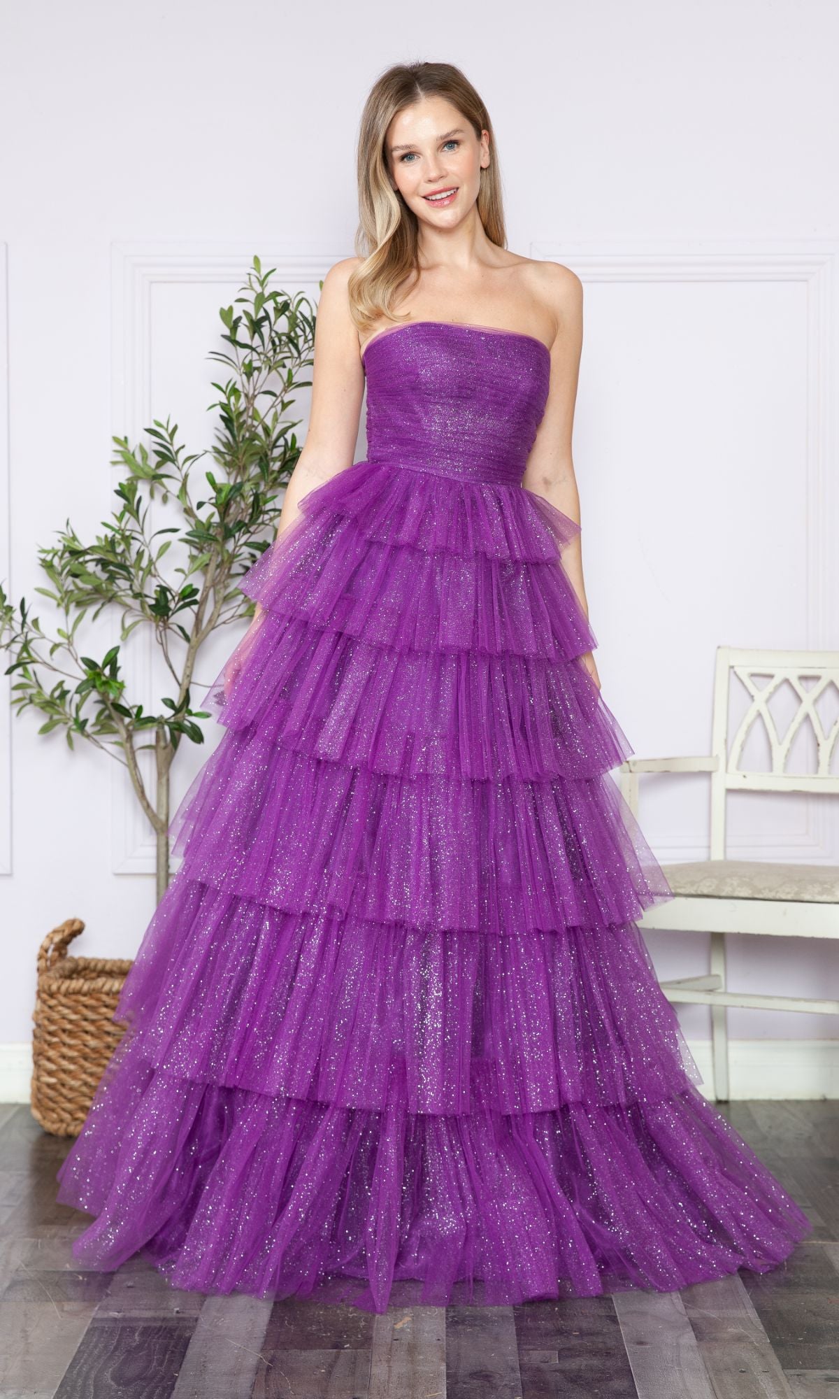 A woman poses indoors wearing the Strapless Long Tiered Glitter Prom Ball Gown 9386 in purple tulle, with ruffles and sparkles. A plant and a white chair are seen in the background.